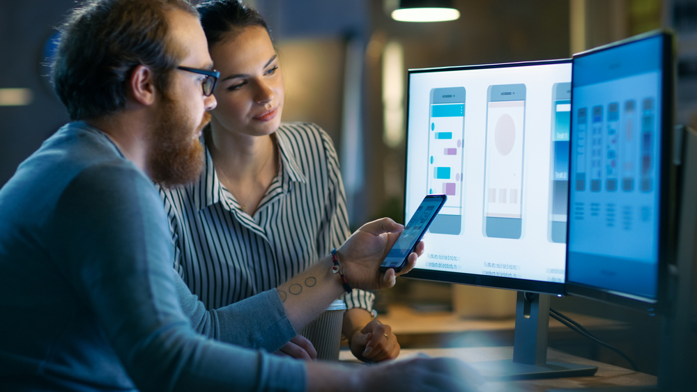 2 App Designers, male, and female, Looking At Mobile App with computer monitors displaying mobile app design coding